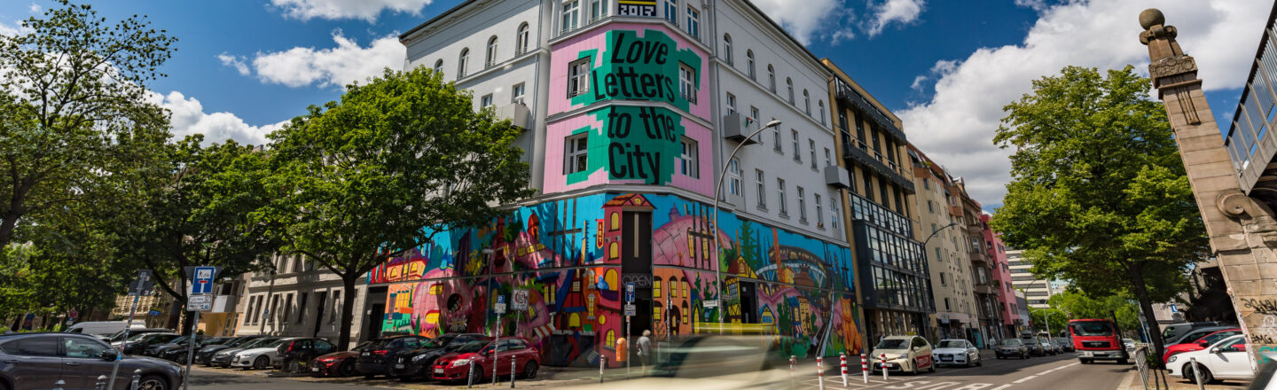 Straßenszene mit einem großen Wandgemälde auf einem Gebäude, das die Worte "Love Letter to the City" zeigt. Das Gemälde ist bunt und zeigt urbane Elemente. Autos sind entlang der Straße geparkt, Bäume säumen den Gehweg, und der Himmel ist teilweise bewölkt.