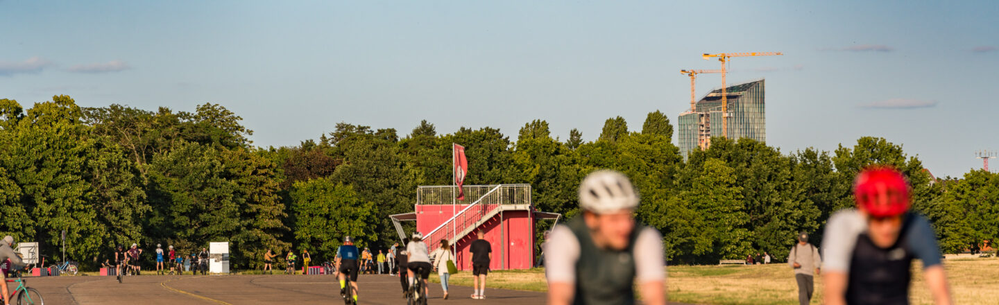 Ein sonniger Tag in einem Park mit Menschen beim Radfahren und Skaten auf einer breiten Asphaltstraße. Im Hintergrund ist ein rotes Gebäude mit einer kleinen Tribüne zu sehen, gefolgt von üppigen grünen Bäumen und einem Baukran neben einem modernen Gebäude. Der Himmel ist blau und leicht bewölkt.
