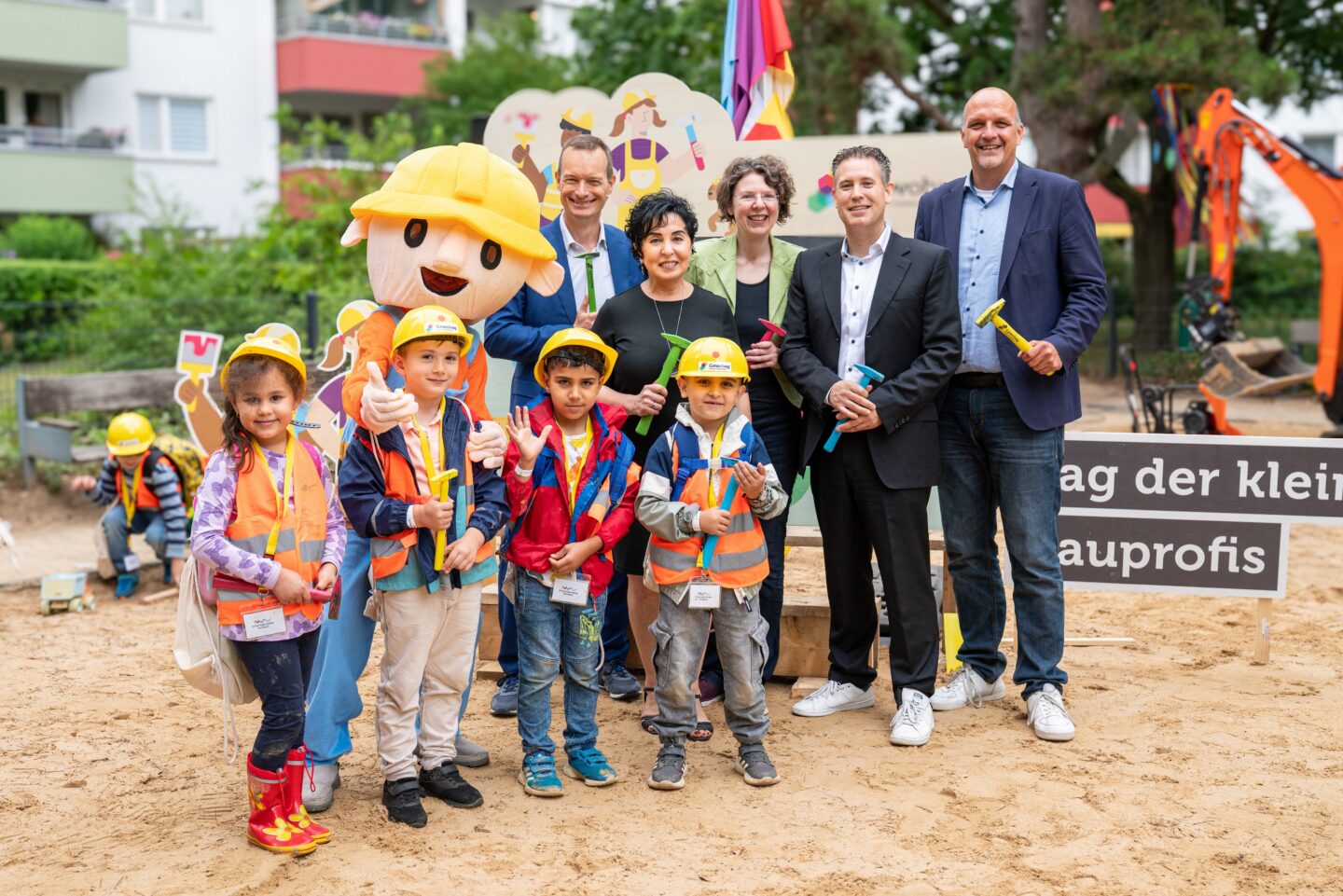 Eine Gruppe von Kindern in Bauarbeiterkostümen, mit Helmen und Sicherheitswesten, steht vor einem Schild mit der Aufschrift "Tag der kleinen Bauprofis". Sie halten bunte Spielwerkzeuge. Neben ihnen steht ein großes Bauarbeiter-Maskottchen. Erwachsene begleiten die Kinder, einige halten ebenfalls Spielzeuge. Im Hintergrund sind Wohngebäude und eine Baggerspielplatzanlage sichtbar.