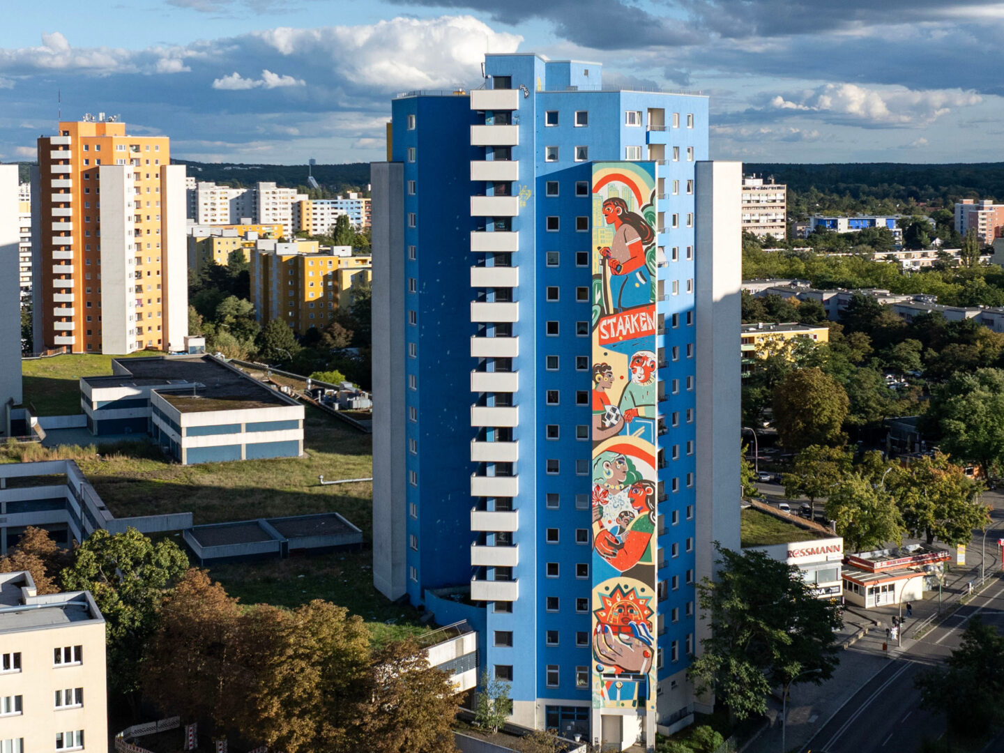Das Bild zeigt ein Hochhaus in einem Wohngebiet. Das Gebäude ist blau gestrichen und hat eine große, farbenfrohe Wandmalerei, die sich über mehrere Stockwerke erstreckt und Figuren sowie den Schriftzug "STAAKEN" zeigt. Rundherum sind weitere Wohnhäuser sichtbar, einige davon in Gelb- und Orangetönen. Die Umgebung ist dicht bebaut, und im Hintergrund sind Bäume und ein bewölkter Himmel zu sehen.