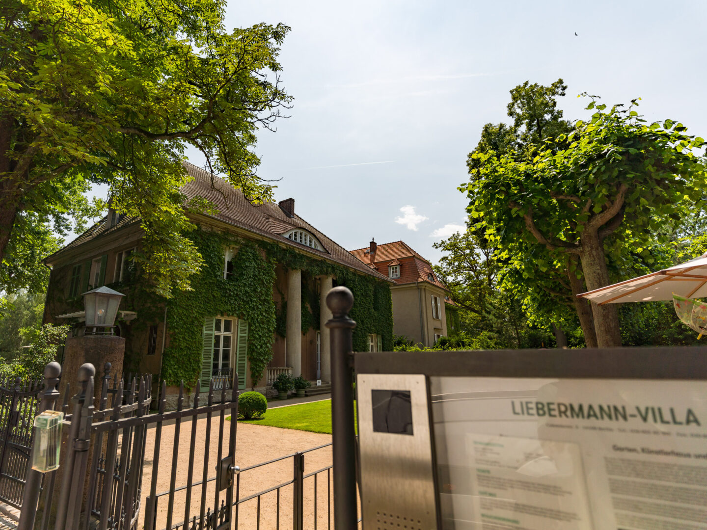 Das Bild zeigt die Liebermann-Villa in Berlin, ein großes Haus mit einer Fassadenfront, die von grünen Ranken und Efeu bewachsen ist. Vor dem Haus befinden sich große Säulen am Eingang. Im Vordergrund ist ein Tor und ein Schild mit der Aufschrift "Liebermann-Villa" sichtbar. Links und rechts vom Haus stehen große, grüne Bäume, und der Himmel ist teils bewölkt mit Sonnenschein.