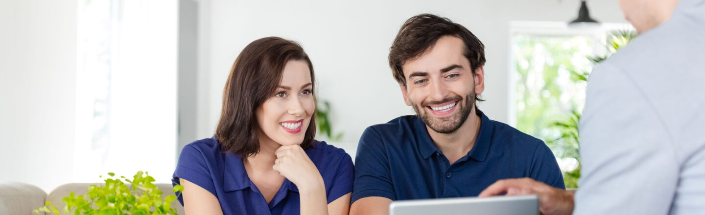 Eine Frau und ein Mann sitzen am Tisch und schauen einen Laptop an, gestikulieren dabei. Im Hintergrund ist grünes Laub sichtbar, das ein frisches Ambiente schafft. Die Szene vermittelt einen Eindruck von Austausch oder Beratung in einer entspannten Umgebung.