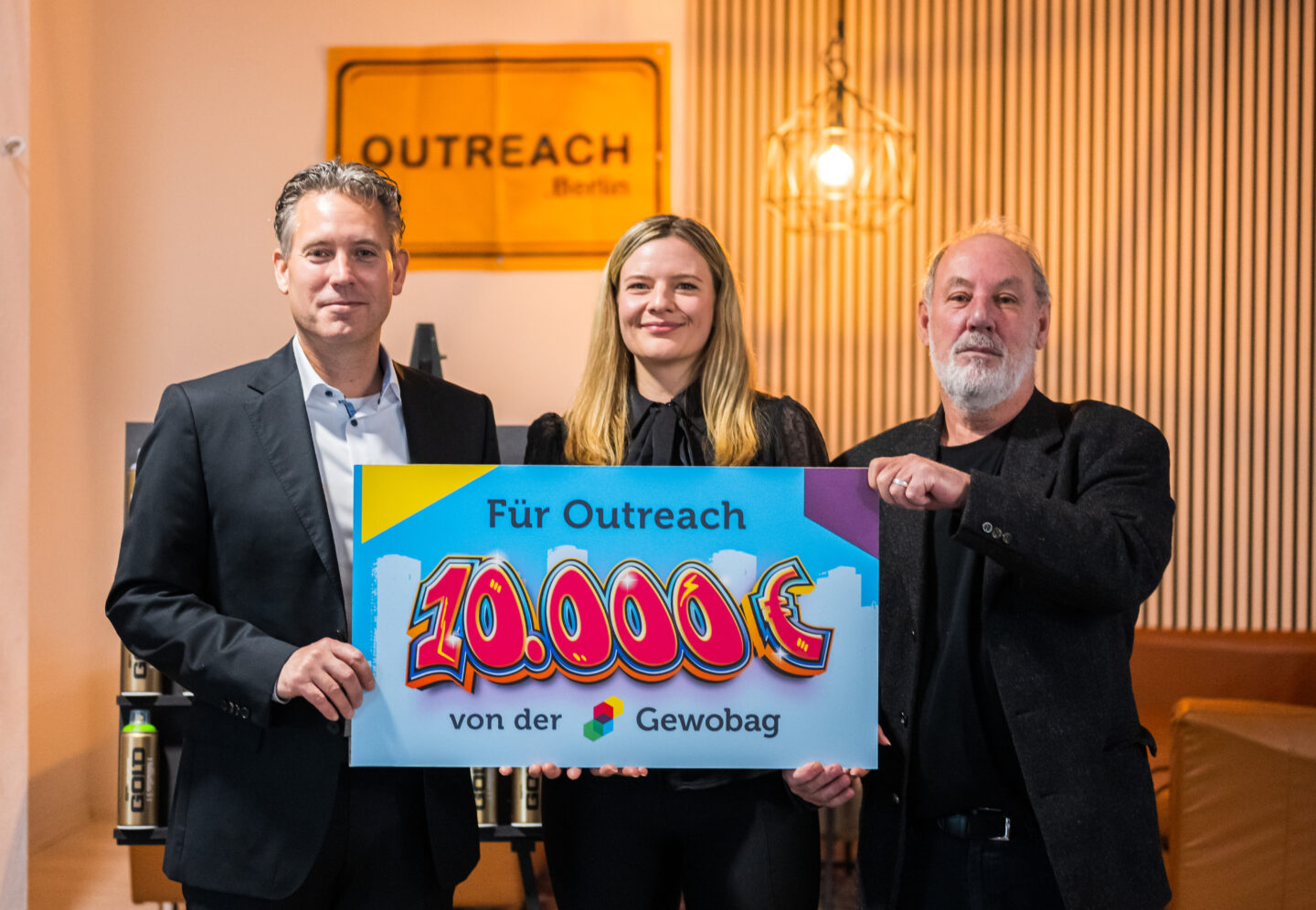 Drei Personen halten gemeinsam ein großes, buntes Schild mit der Aufschrift "Für Outreach 10.000 € von der Gewobag". Im Hintergrund ist ein Schild mit der Aufschrift "OUTREACH Berlin" zu sehen, sowie dekorative Beleuchtung und vertikale Streifen an der Wand.