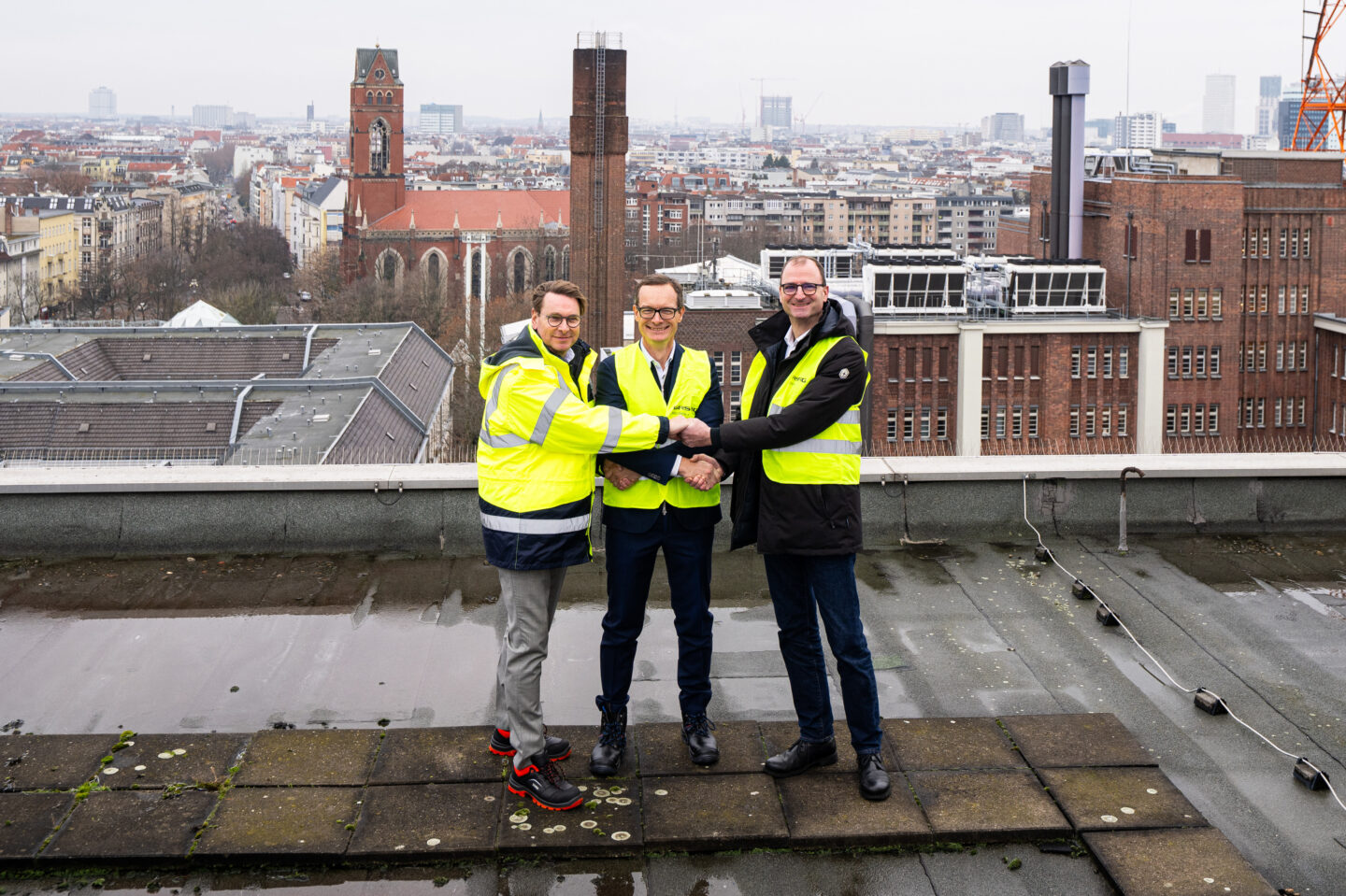 Drei Personen in gelben Sicherheitswesten stehen auf einem Dach mit einer Stadtlandschaft im Hintergrund, die Gebäude und eine Kirche zeigt. Sie geben sich die Hände, um eine Vereinbarung oder Zusammenarbeit zu symbolisieren.