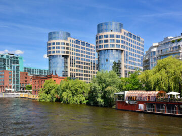 Moderne Gebäude aus Glas und Stein am Flussufer, umgeben von Bäumen. Ein Restaurantboot mit verglaster Terrasse im Vordergrund.