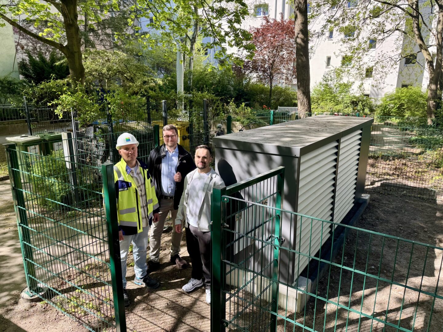 3 Personen stehen vor einer großen Wärmepumpe in der Außenanlage einer Wohnsiedlung.