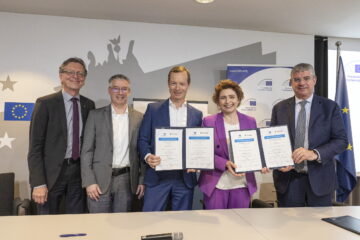 Fünf Personen stehen bei einer EU-Veranstaltung und präsentieren unterschriebene Vertragsdokumente in einem Konferenzraum.