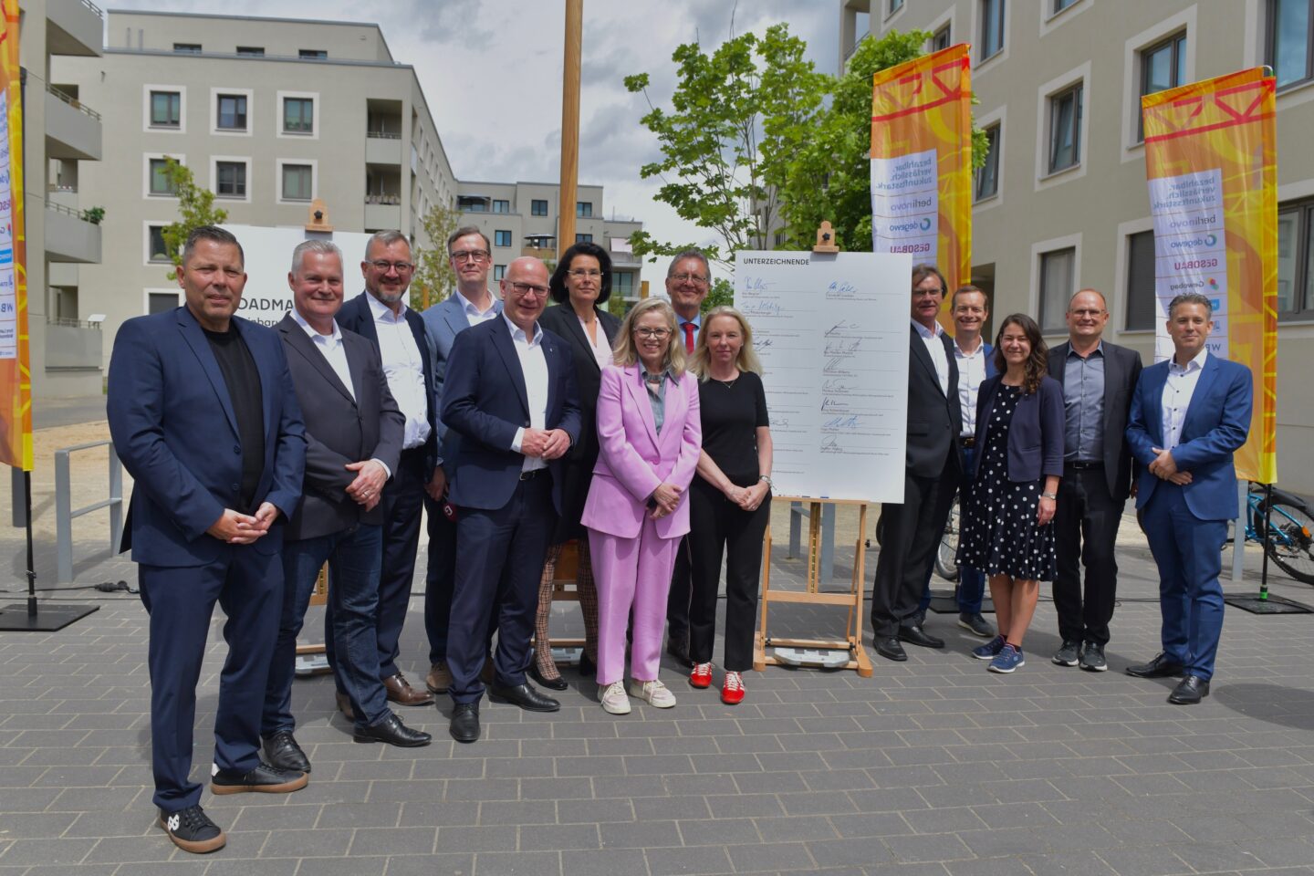 Gruppe von Geschäftsleuten steht im Freien vor Wohngebäuden um ein großes unterschriebenes Plakat, Banner und Bäume im Hintergrund.