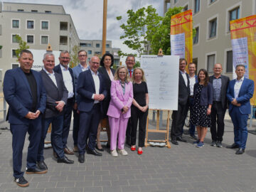 Gruppe von Geschäftsleuten steht im Freien vor Wohngebäuden um ein großes unterschriebenes Plakat, Banner und Bäume im Hintergrund.