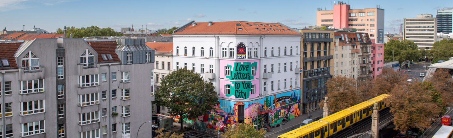 Bunte Hausfassade mit Graffiti „Love letters to the City“ an Berliner Straße, davor fährt ein gelber U‑Bahnzug auf Hochgleis vorbei.