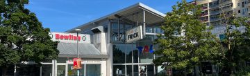 Eingang eines Einkaufs- und Freizeitcenters mit Bowling-Aufschrift und großem „Frick“-Schild, umgeben von Bäumen und Hochhäusern.