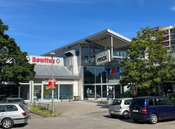 Einkaufs- und Freizeitcenter mit Bowling-Aufschrift und großem „Frick“-Schild, davor mehrere parkende Autos und Bäume bei Sonnenschein.