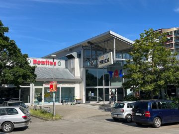 Einkaufs- und Freizeitcenter mit Bowling-Aufschrift und großem „Frick“-Schild, davor mehrere parkende Autos und Bäume bei Sonnenschein.