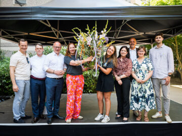 Gruppe von neun Erwachsenen steht lächelnd auf einer Bühne im Freien, zwei Frauen in der Mitte halten einen großen Blumenstrauß mit bunten Bändern.