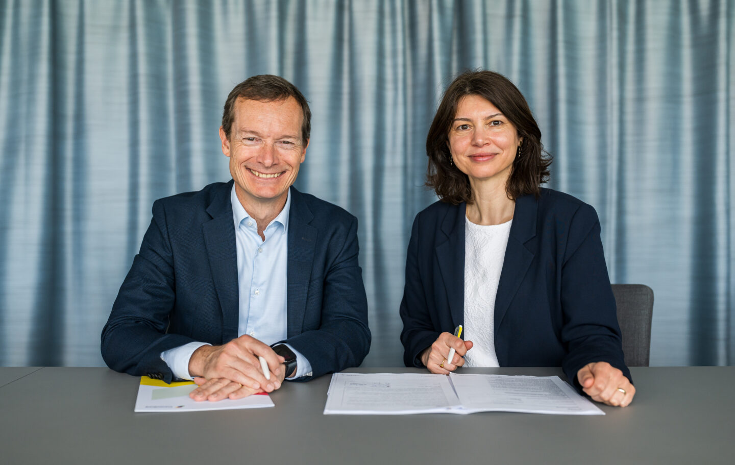 Zwei Personen in Businesskleidung sitzen am Tisch vor Vertragsunterlagen, halten Stifte in der Hand, Hintergrund ist ein grauer Vorhang.