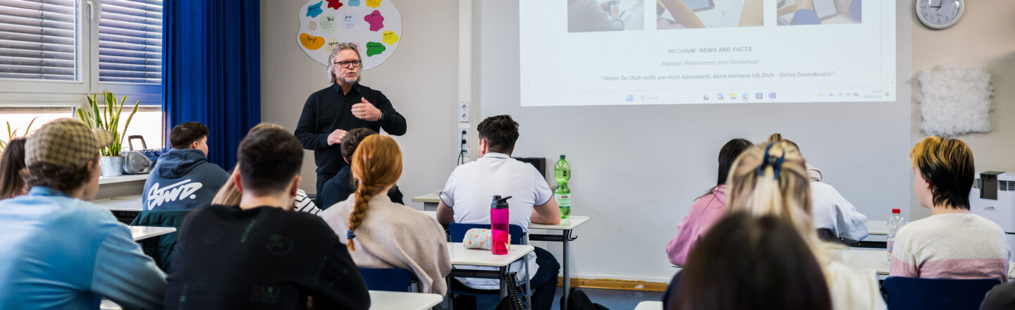 Schulklasse sitzt an Tischen und folgt einem Vortragenden vor einer projizierten Präsentation; Blick von hinten auf die Schülerinnen und Schüler.