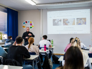 Schulklasse sitzt an Tischen und folgt einem Vortragenden vor einer projizierten Präsentation; Blick von hinten auf die Schülerinnen und Schüler.