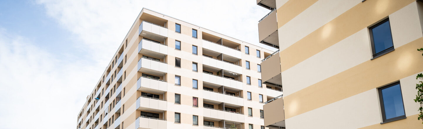 Helle, mehrstöckige Wohnhäuser mit Balkonen vor leicht bewölktem Himmel, moderne Fassaden in Beige- und Weißtönen, Blick nach oben.