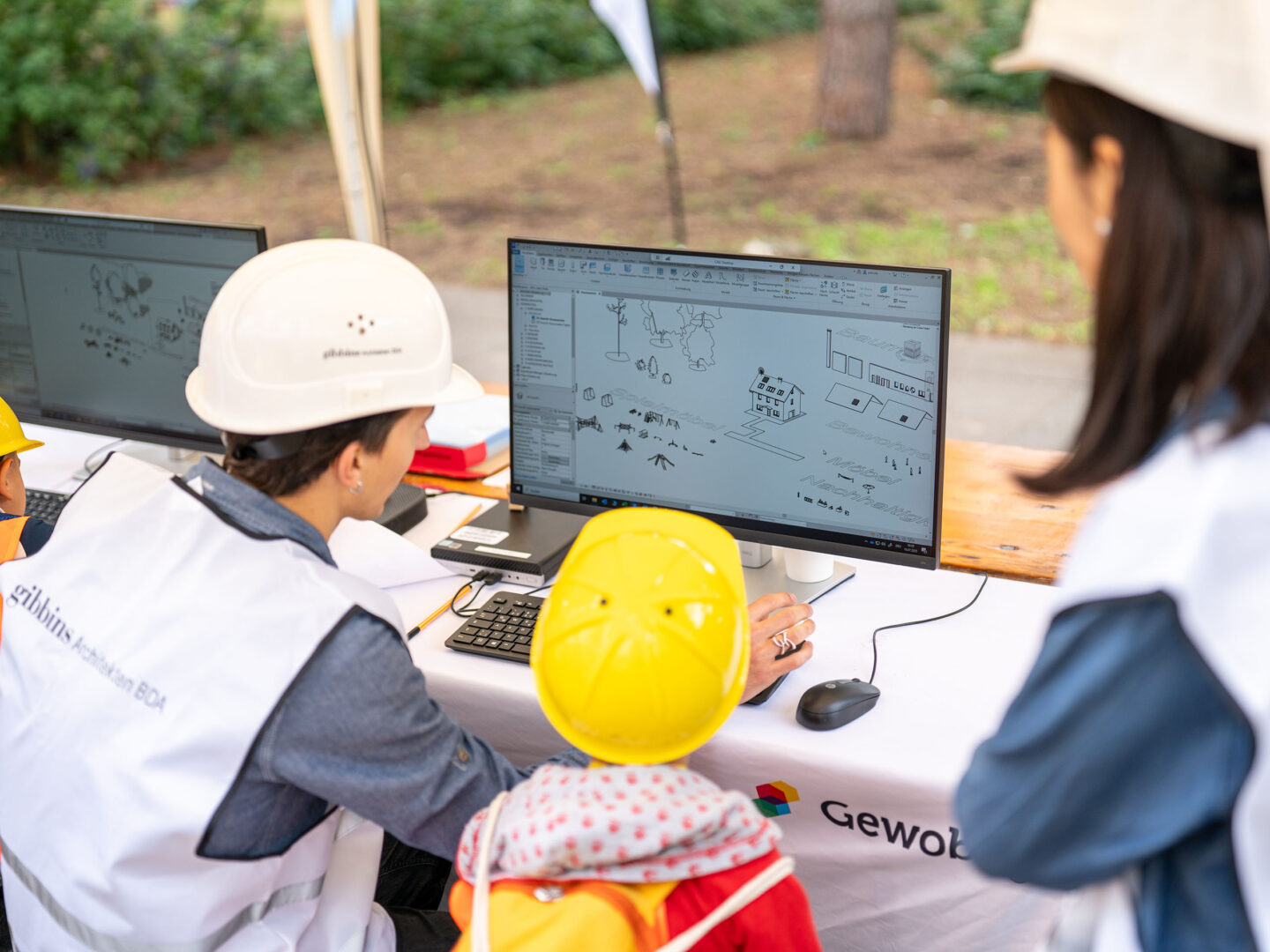 01 Kinder mit Bauhelmen schauen einem Planer am Computer zu, der am Bildschirm ein Bauprojekt mit Häusern, Wegen und Spielplatz zeichnet. | Fotos: Ralph Maak