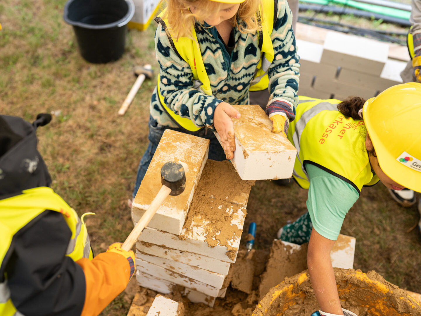 Kinder mit Bauhelmen und Warnwesten mauern spielerisch: Sie stapeln sandige Ziegelsteine und arbeiten gemeinsam auf der Wiese.