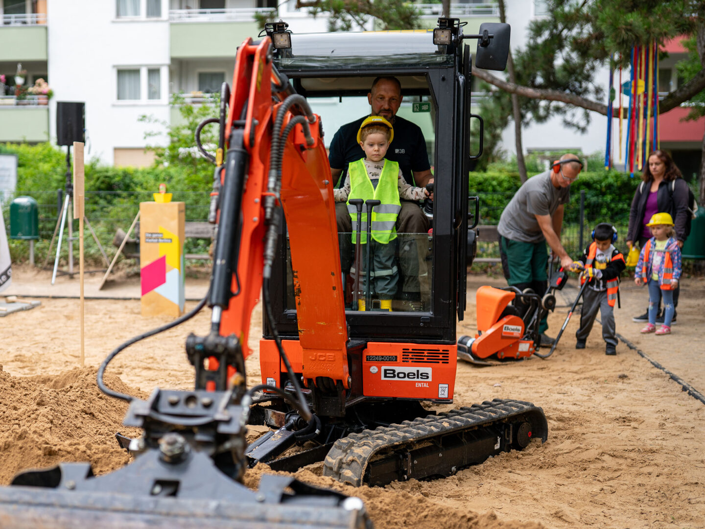 Kind im Bauarbeiter-Outfit sitzt mit Erwachsenen im Minibagger, andere Kinder erkunden spielerisch die Baustelle im Sand.