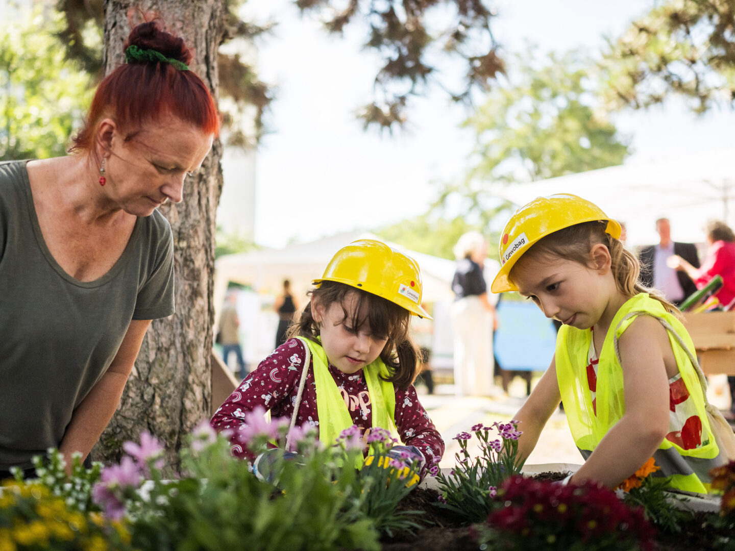 Frau und zwei Kinder mit gelben Bauhelmen pflanzen gemeinsam bunte Blumen im Garten; die Kinder tragen Warnwesten und arbeiten konzentriert.