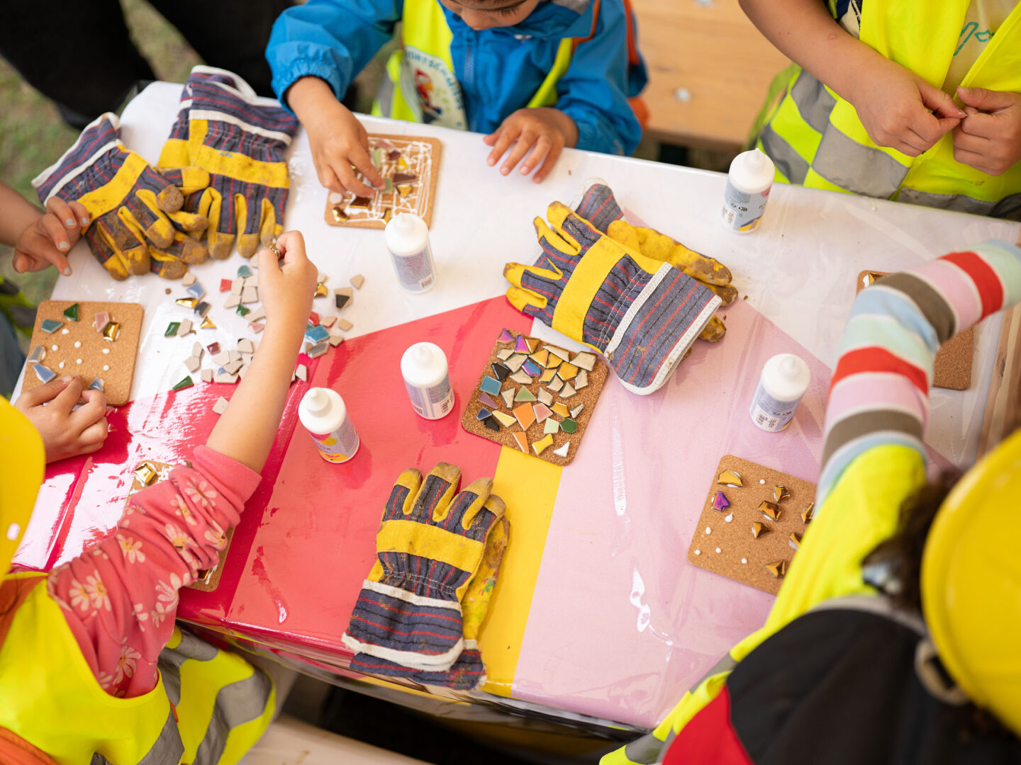 Kinder basteln am Tisch bunte Mosaike auf Kacheln; Bauhandschuhe und Kleber liegen bereit, alle tragen Sicherheitswesten.
