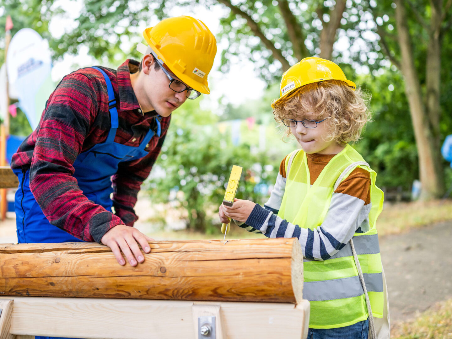 Erwachsener und Kind mit Schutzhelmen arbeiten draußen an einem Holzstamm; das Kind schlägt einen Nagel ein, der Erwachsene unterstützt.