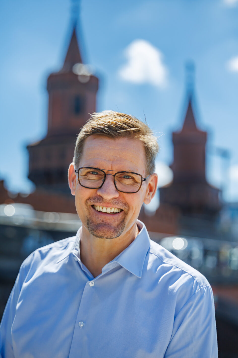 Porträt eines lächelnden Mannes mit Brille im blauen Hemd vor verschwommenen Backsteintürmen unter blauem Himmel mit Wolken.