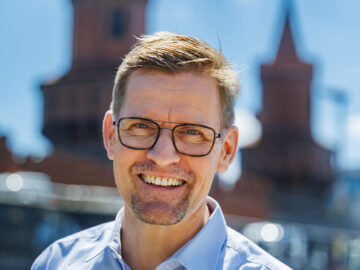 Porträt eines lächelnden Mannes mit Brille im blauen Hemd vor verschwommenen Backsteintürmen unter blauem Himmel mit Wolken.
