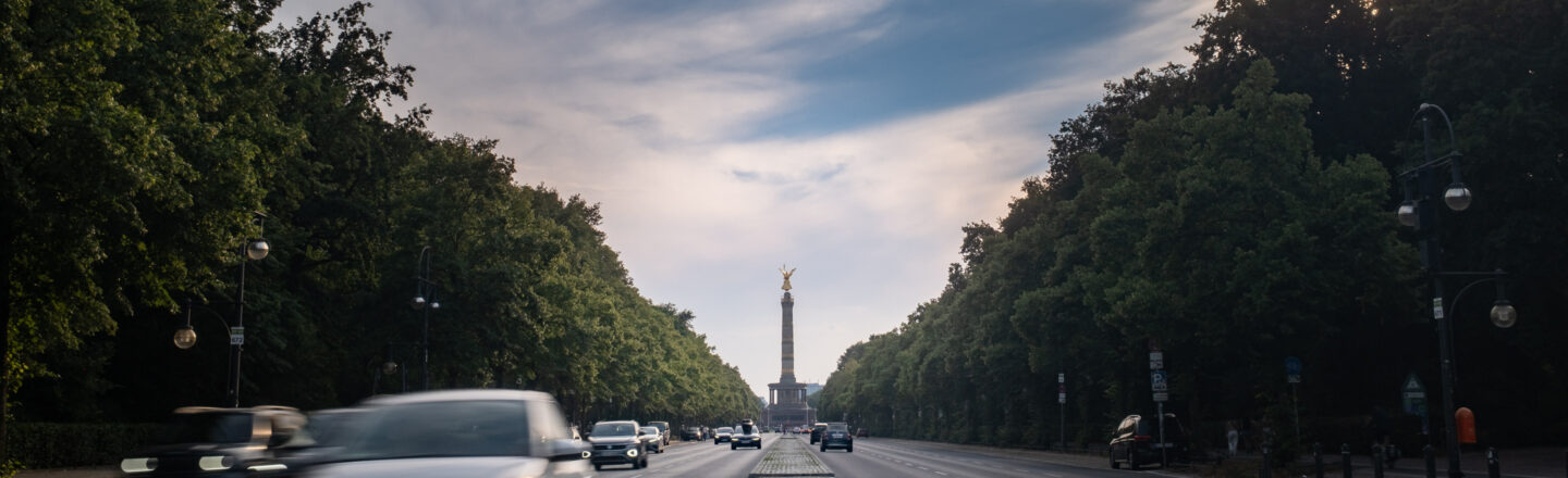 Berliner Straße im Tiergarten mit Autos, gesäumt von dichtem Grün; am Horizont steht die Siegessäule unter bewölktem Himmel.