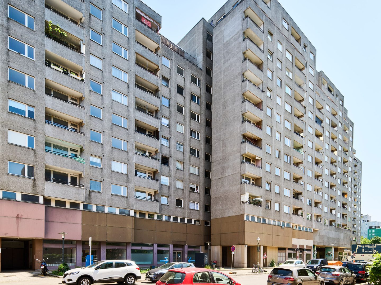 Straßenszene mit hohen grauen Wohnblocks mit vielen Balkonen; davor parken zahlreiche Autos am sonnigen Tag.