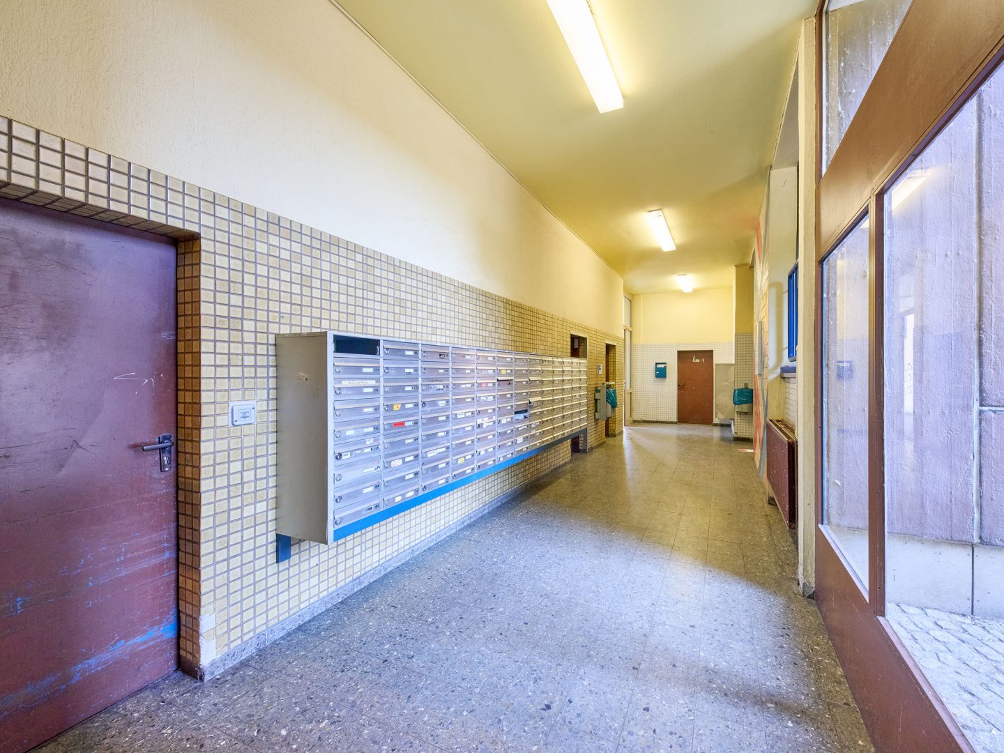 Langer, schlicht beleuchteter Hausflur mit vielen silbernen Briefkästen an der Wand, mehreren Wohnungstüren und großen Fenstern rechts.
