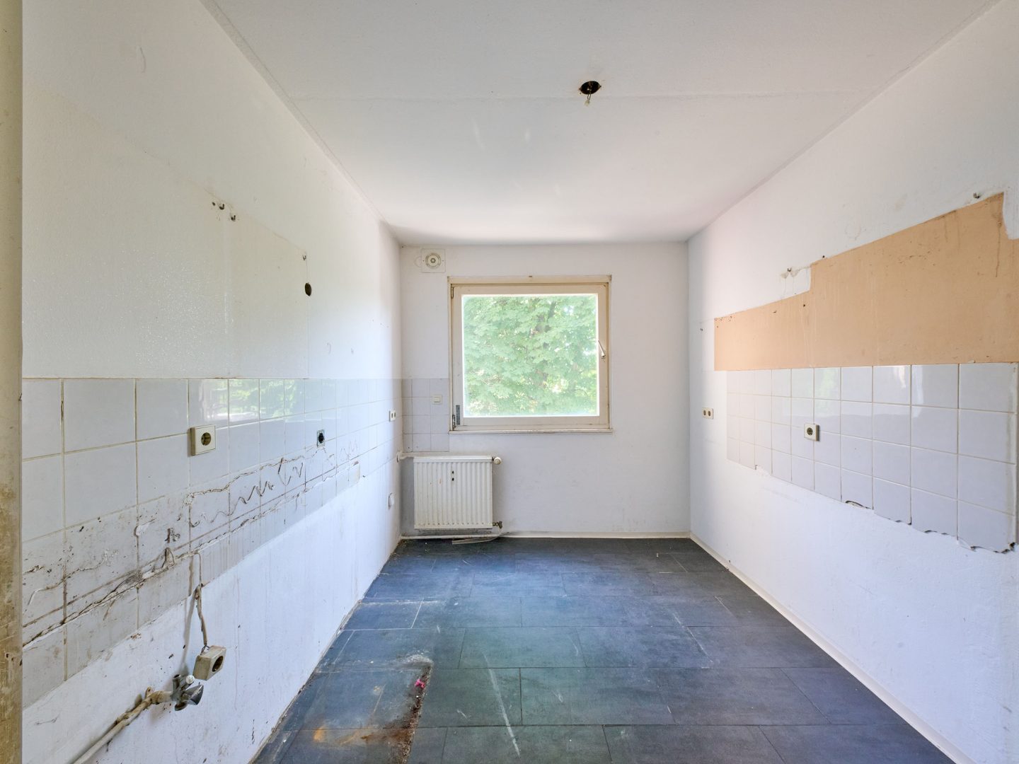 Leerer, unsanierter Küchenraum mit abmontierten Einbauten, sichtbaren Leitungen, weißen Fliesen und Fenster mit Blick ins Grüne.