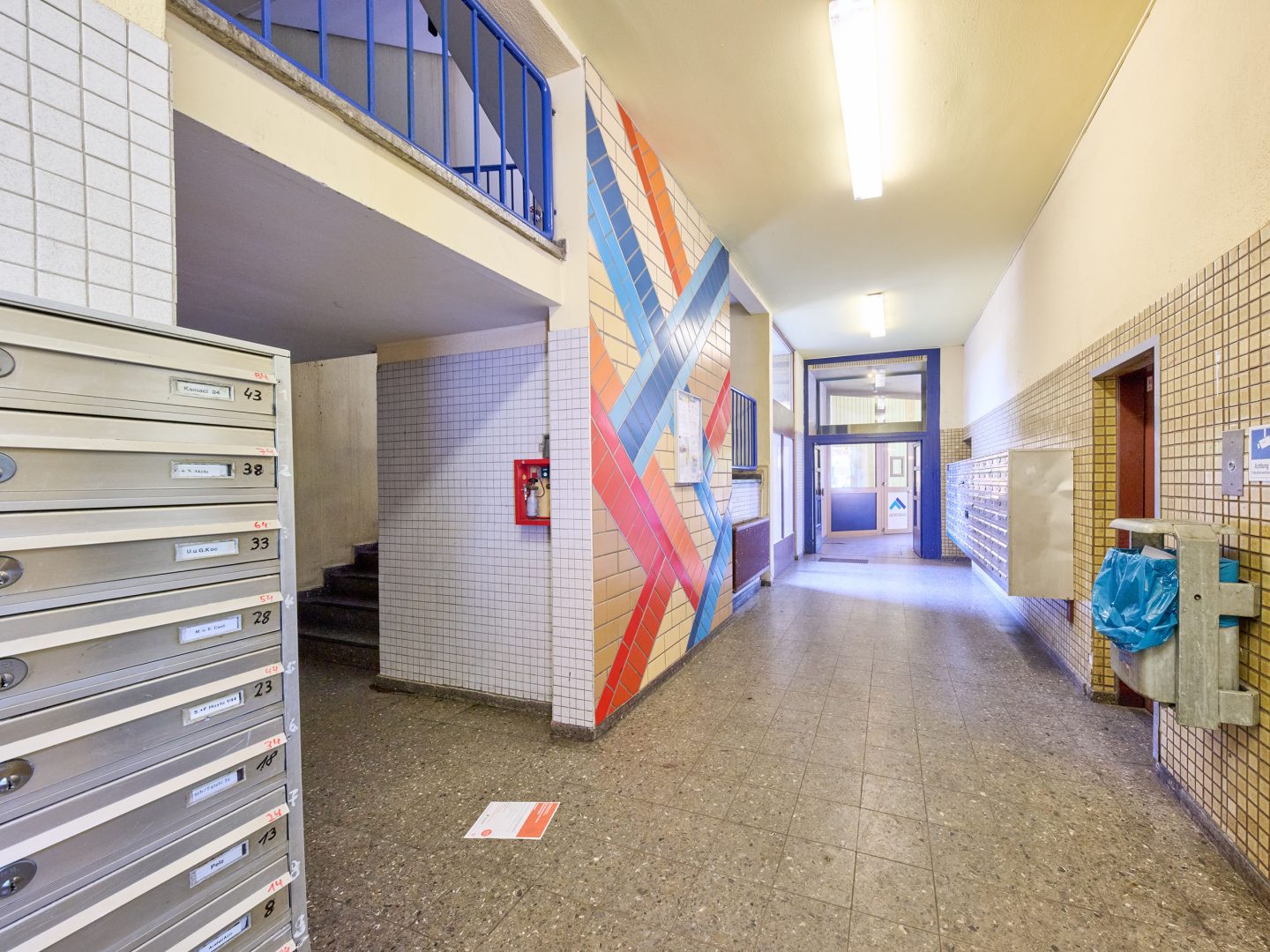 Hausflur mit Briefkästen, Treppenaufgang und bunter Wandmalerei aus Kreuzstreifen; am Ende Glas-Eingangstür, rechts Papiermüllbehälter.
