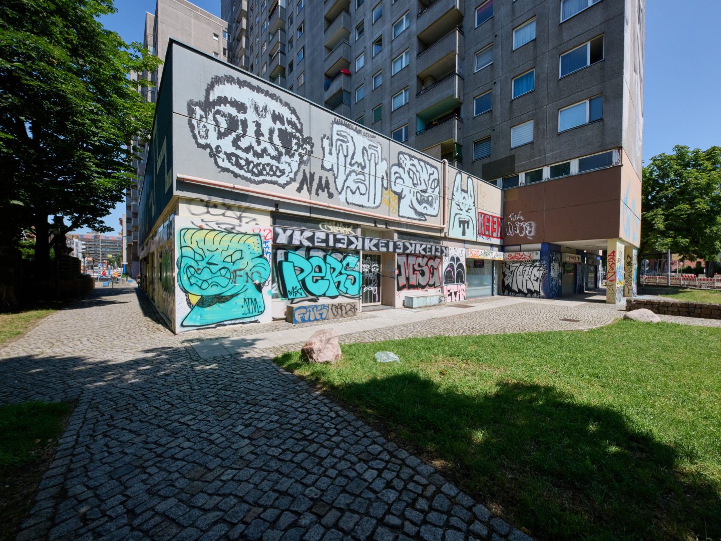 Vor einem grauen Hochhaus steht ein flacher Anbau, dessen Fassaden großflächig mit bunten Graffiti besprüht sind; davor Weg und Rasen.