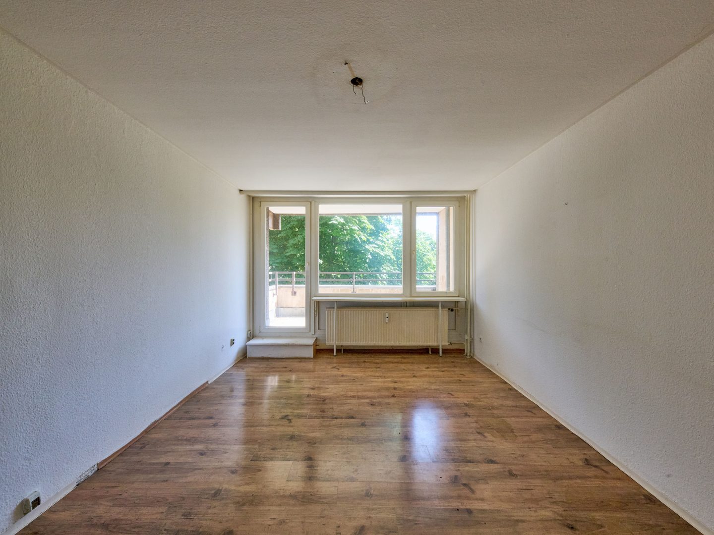 Leeres, längliches Wohnzimmer mit Holzfußboden, großer Fensterfront zum Balkon und Blick auf grüne Bäume.