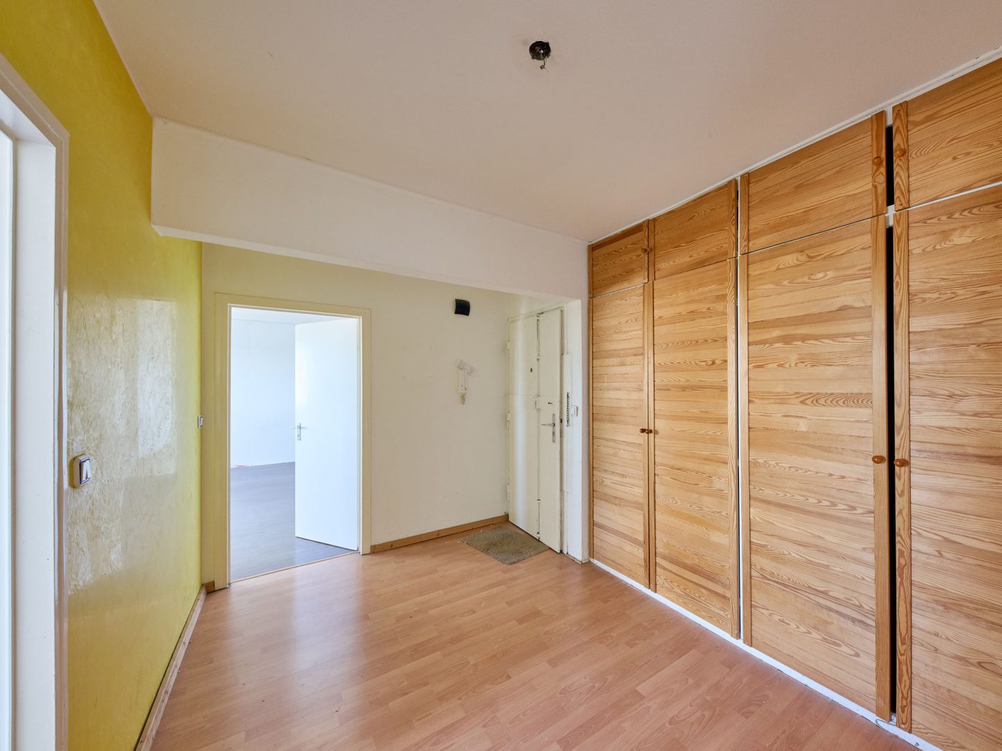 Leerer Flur mit Laminatboden, großer Einbauschrank aus Holz rechts, gelb gestrichene Wand links und offener Tür in ein helles Zimmer.