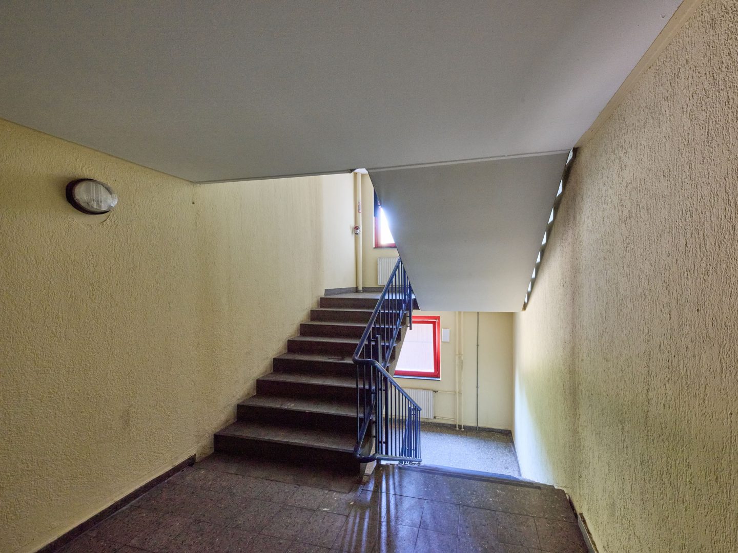 Einfaches Treppenhaus mit gelb gestrichenen Wänden, dunklen Stufen und blauem Geländer, am Ende ein Fenster mit Tageslicht.