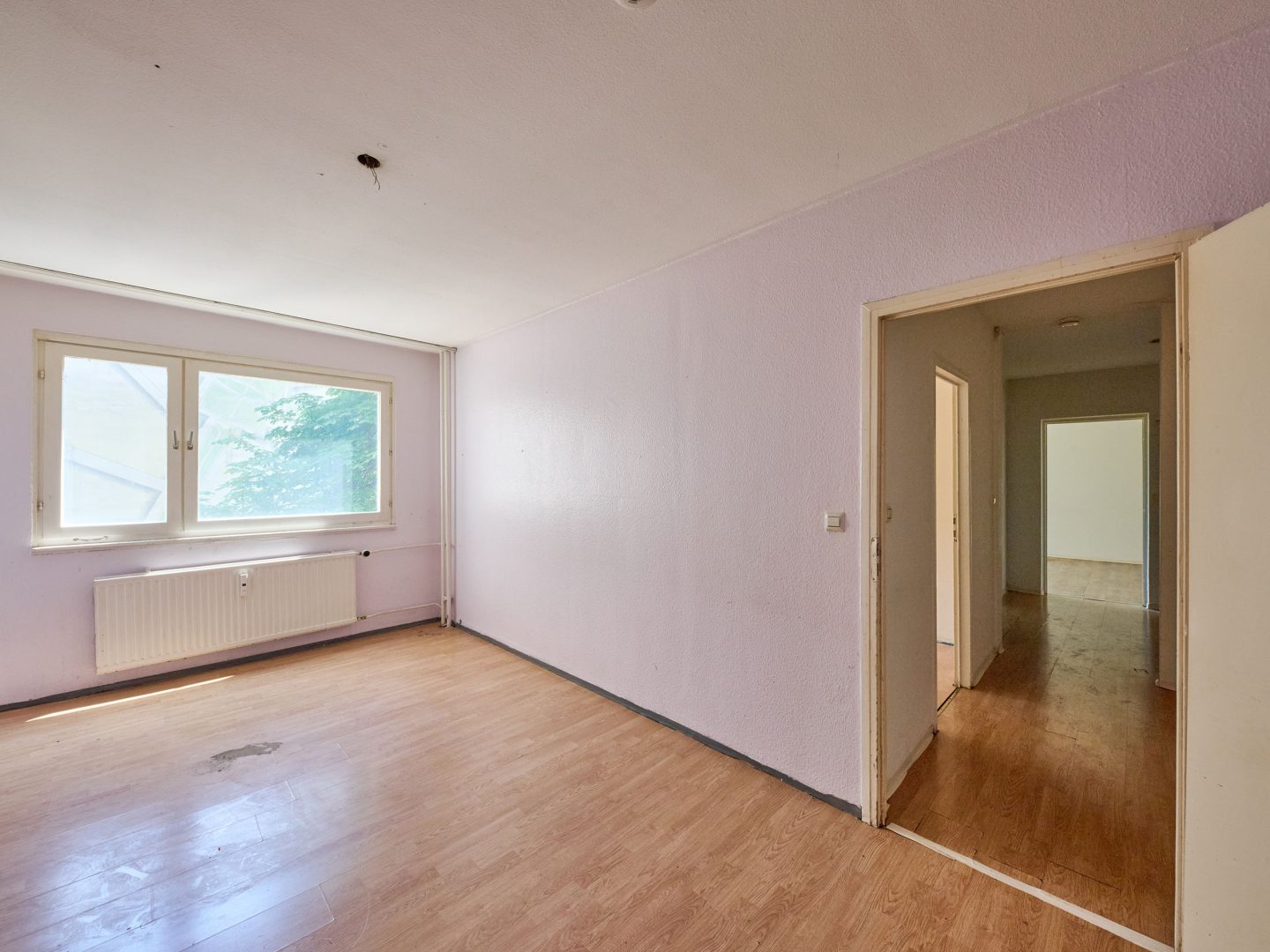 Leeres Zimmer mit Laminatboden und helllila Wänden, links Fenster mit Blick ins Grüne, rechts offene Tür zum Flur mit weiteren Räumen.