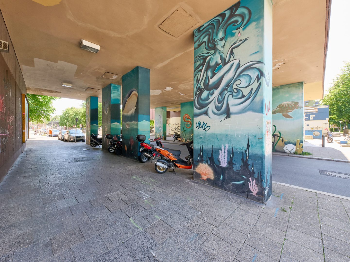 Unter einem Wohnblock stehen Motorroller; mehrere Pfeiler des Durchgangs sind bunt mit Unterwasser‑ und Figurenmotiven bemalt.