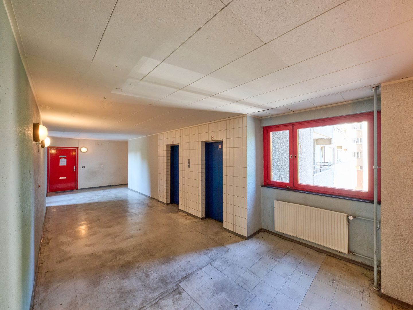 Heller Flur mit zwei blauen Aufzügen und rotem Fenster, links eine rote Tür; Betonboden, Beleuchtung an den Wänden.