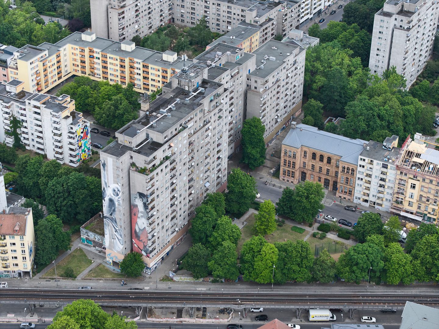 Luftaufnahme grauer Wohnblocks mit großem Fassadenkunstwerk, umgeben von Bäumen, weiteren Häusern und unten verlaufenden Bahngleisen.