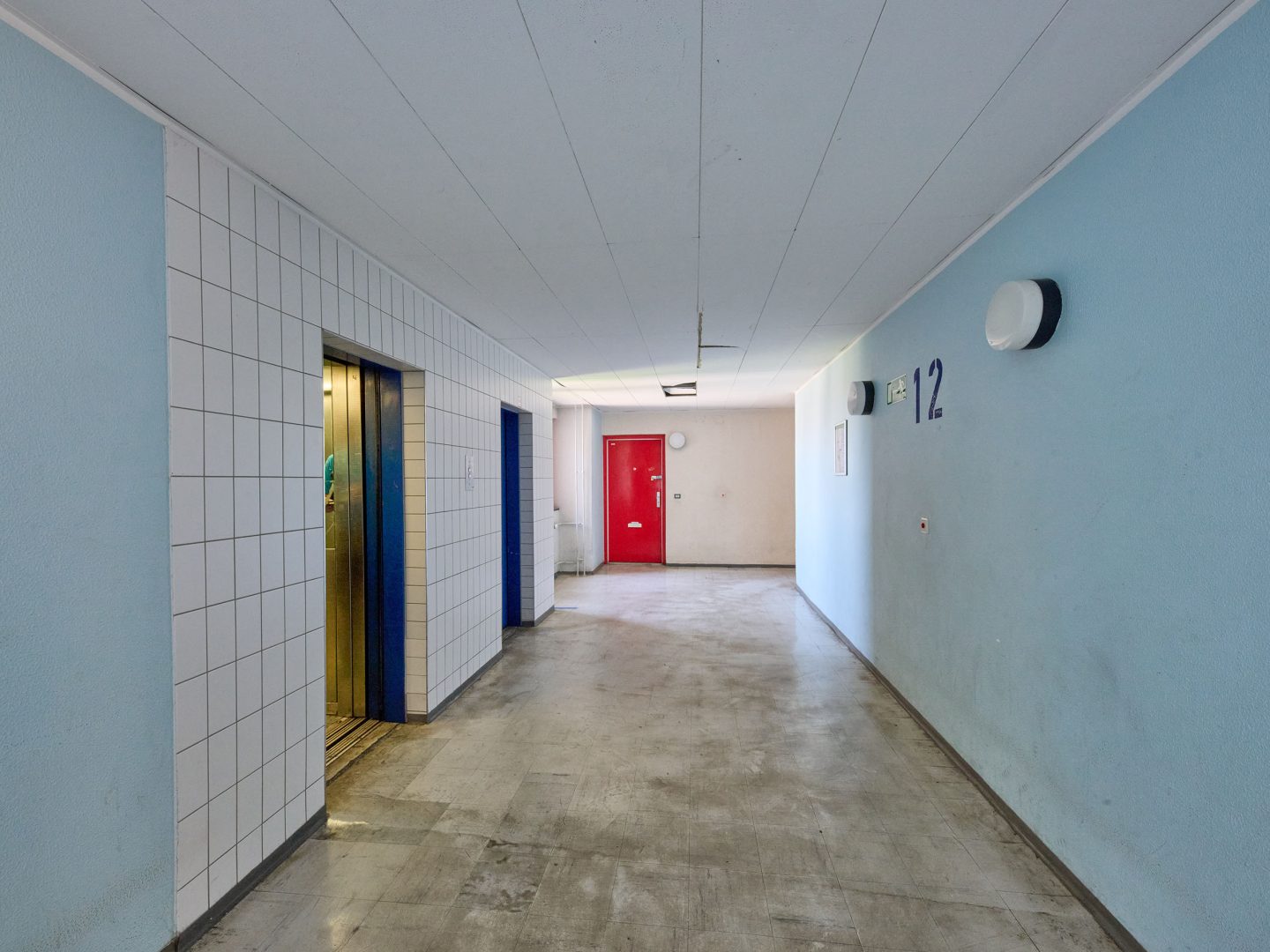 Langer Hausflur im 12. Stock mit blauen Aufzugtüren links und roter Wohnungstür am Ende, grauer Boden und hellblauen Wänden.