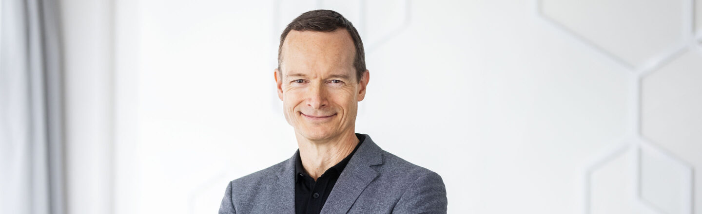 Mann im grauen Sakko steht mit verschränkten Armen vor heller Wand mit Gewobag‑Logo und lächelt in die Kamera.