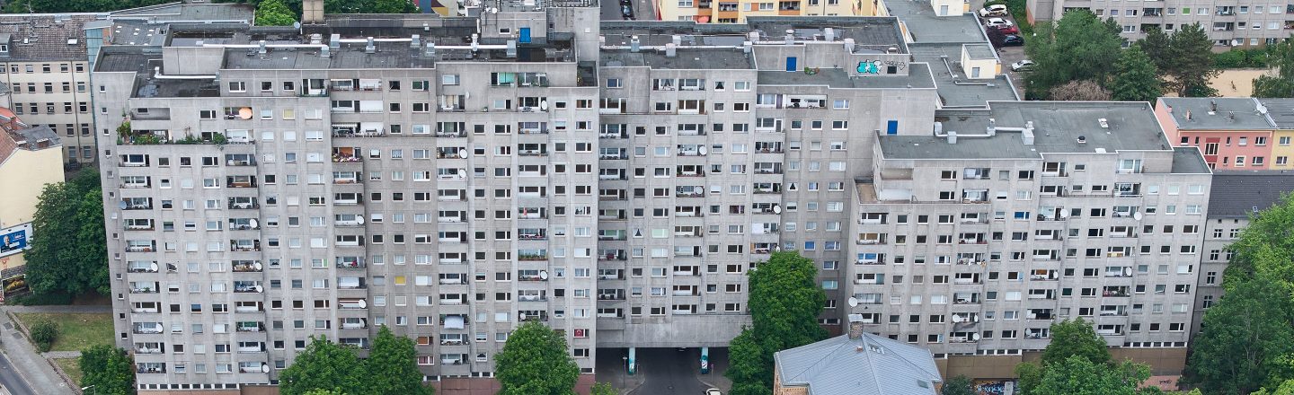 Luftaufnahme eines langen grauen Plattenbaus mit vielen Balkonen und Fenstern, umgeben von Bäumen und angrenzenden Wohnhäusern.