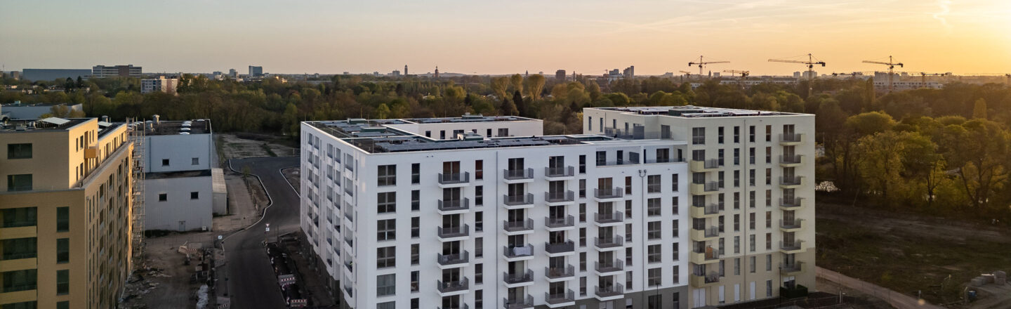 Luftaufnahme eines neuen weißen Wohnblocks mit Balkonen in einem Baugebiet, umgeben von Baustellen und Bäumen im Abendlicht.