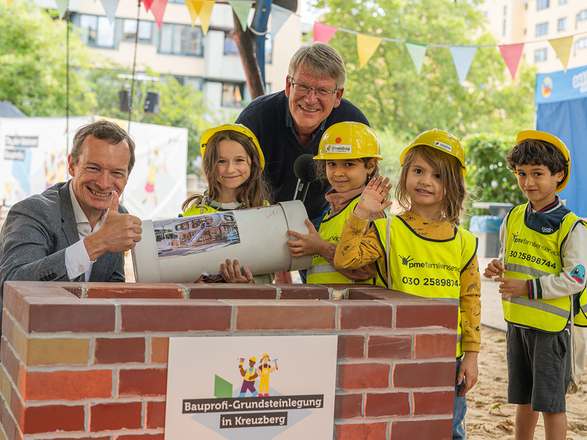 Kinder mit Bauhelmen und Warnwesten legen mit zwei Erwachsenen den Grundstein, alle lächeln hinter einer Mauer mit Baustellenschild.