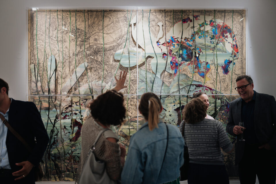 Menschen stehen in einer Galerie vor einem großen, bunten Wandbild und diskutieren angeregt über das Kunstwerk.