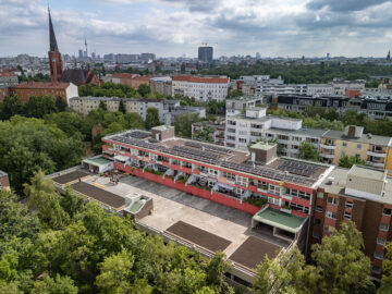 Luftaufnahme eines Wohnblocks mit Solaranlagen auf dem Dach; auf dem Vordach bereiten Menschen lange Beete für einen Gemeinschaftsgarten vor.